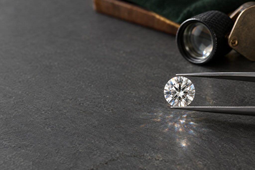 Loose colourless round brilliant diamond held in tweezers on grey slate beside a jeweller's loupe