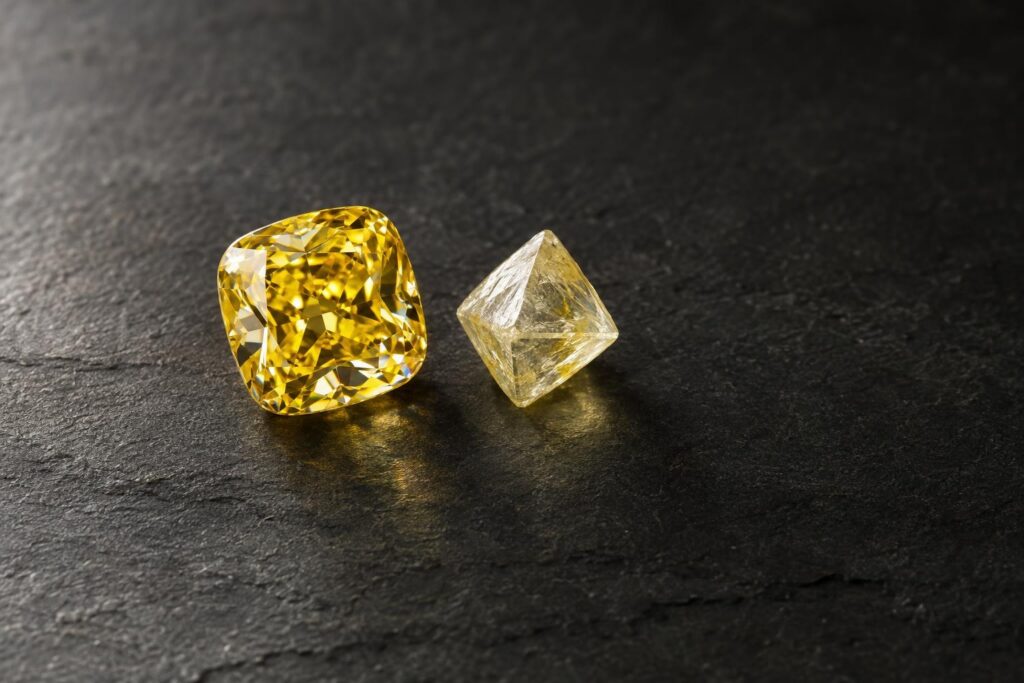 Yellow cushion-cut diamond beside a rough yellowish octahedral diamond crystal on slate
