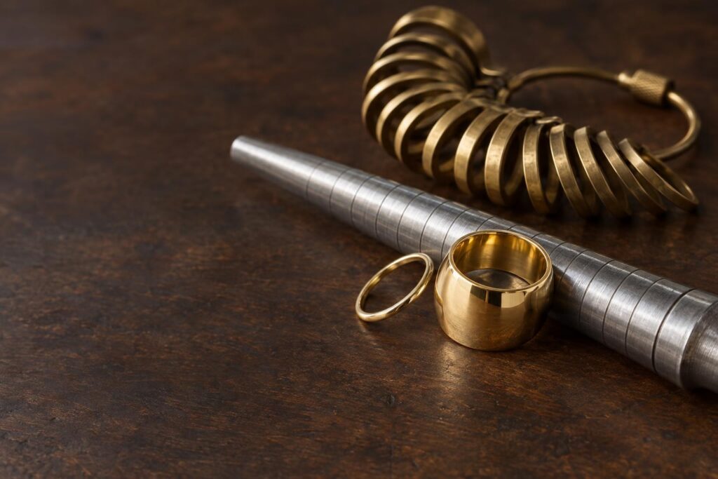 Wide and thin gold bands beside a ring mandrel and finger sizing gauge on a jeweller's bench